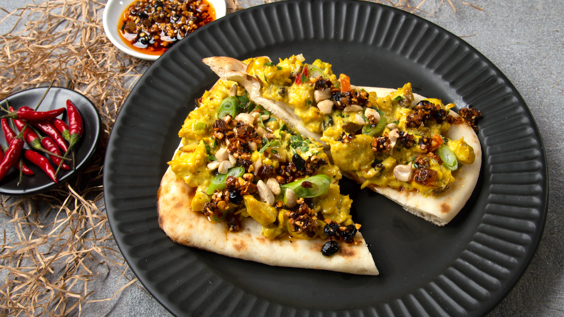 Black plate with Indian flatbread and curried chicken salad topped with oo’mämē Chinese Chile infusion. Small black bowl of red chilis. #myoomame