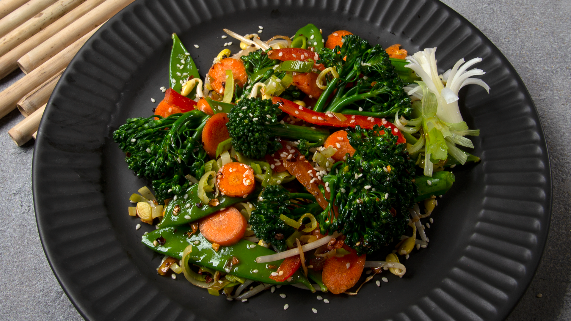 Black plate with veggie stir fry and spicy oo’mämē, broccolini, peppers, carrots, snow peans and bean shoots with hemp hearts #myoomame
