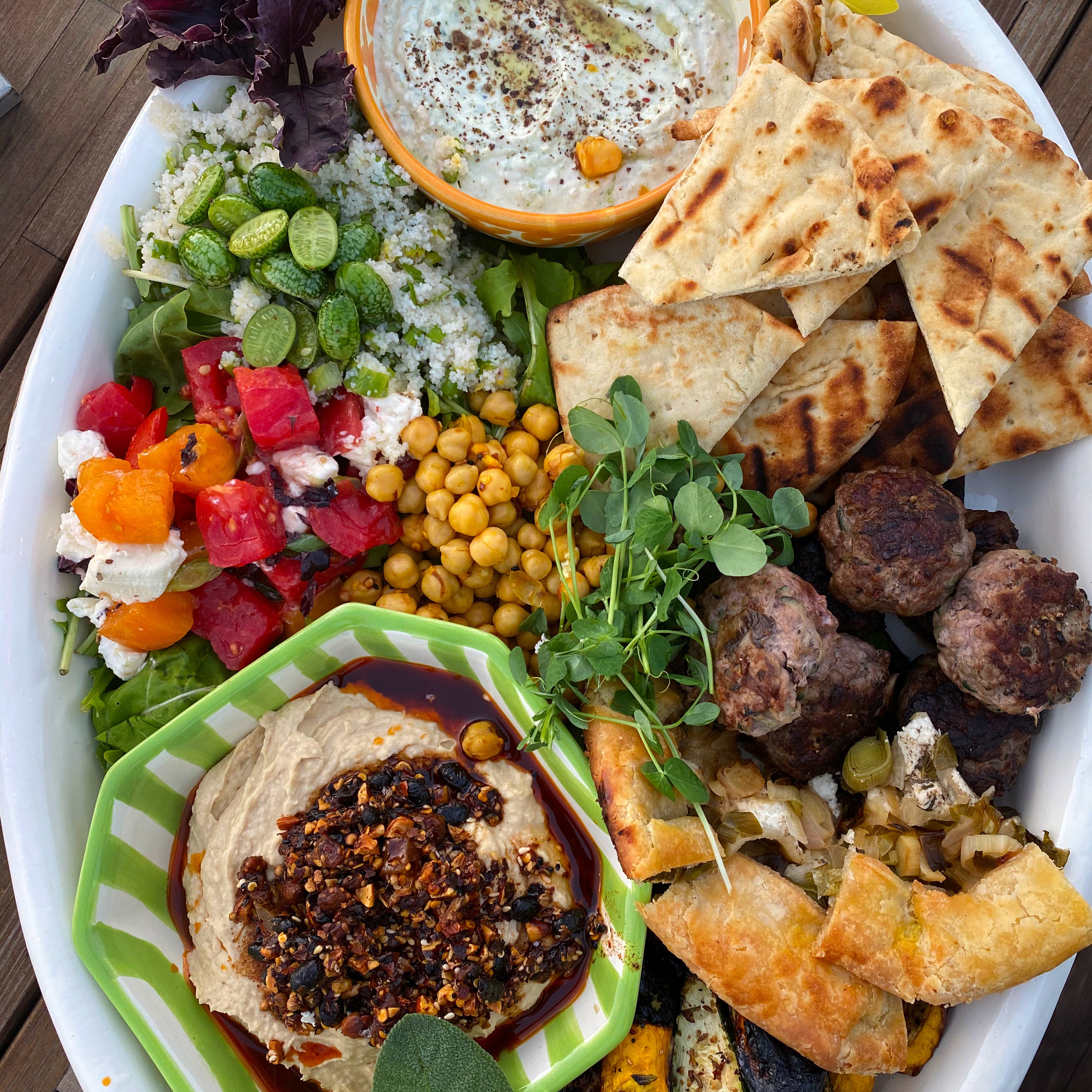 Large white oval platter with oo’mämē lamb sliders, leek galette, grilled squash, hummos topped with oo’mämē, tomato and sheep cheese, fonio with baby cucumbers, garbanzos, naan wedges, yogurt dip with spices and herb garnish.