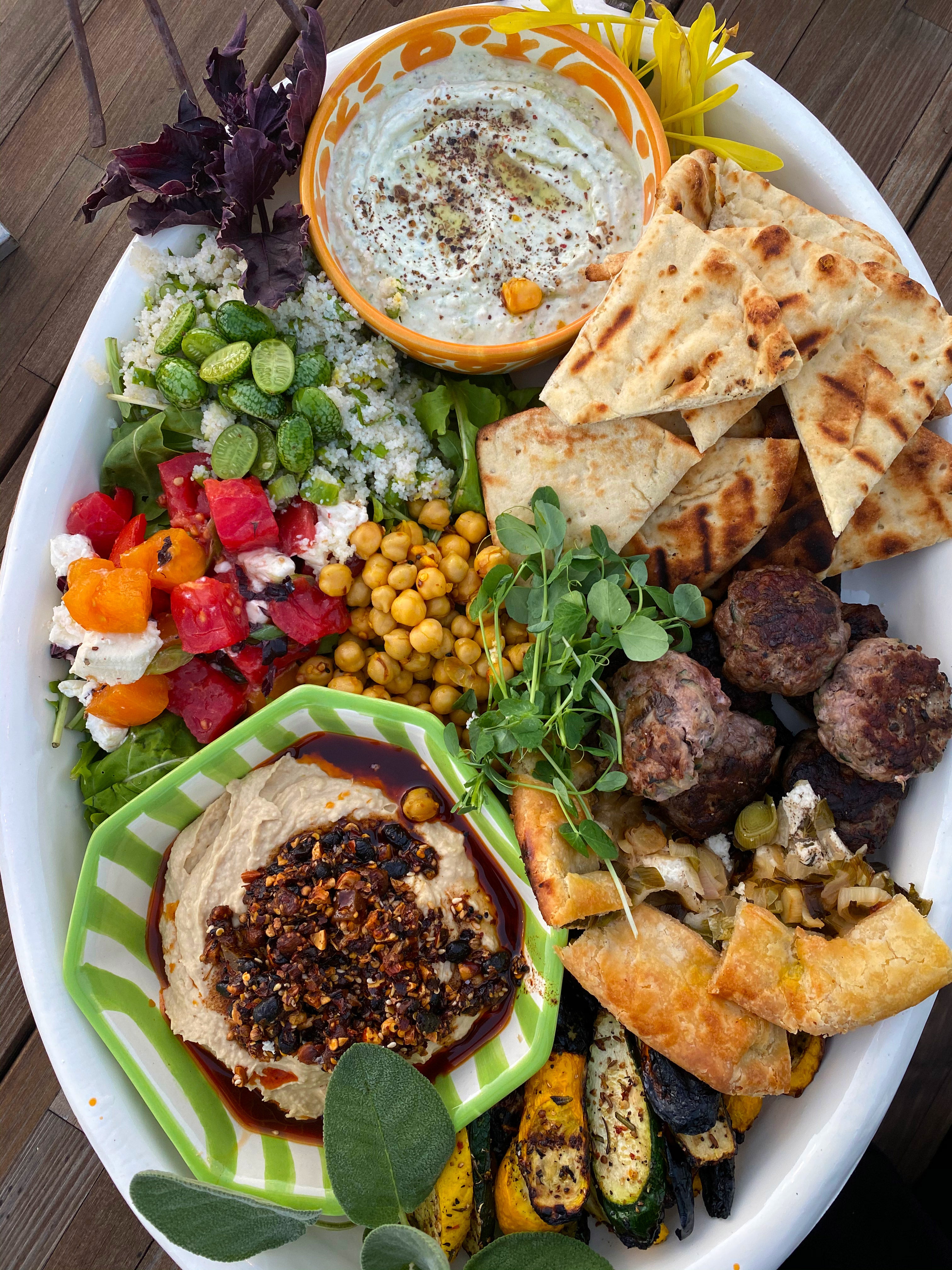 Large white oval platter with oo’mämē lamb sliders, leek galette, grilled squash, hummos topped with oo’mämē, tomato and sheep cheese, fonio with baby cucumbers, garbanzos, naan wedges, yogurt dip with spices and herb garnish.