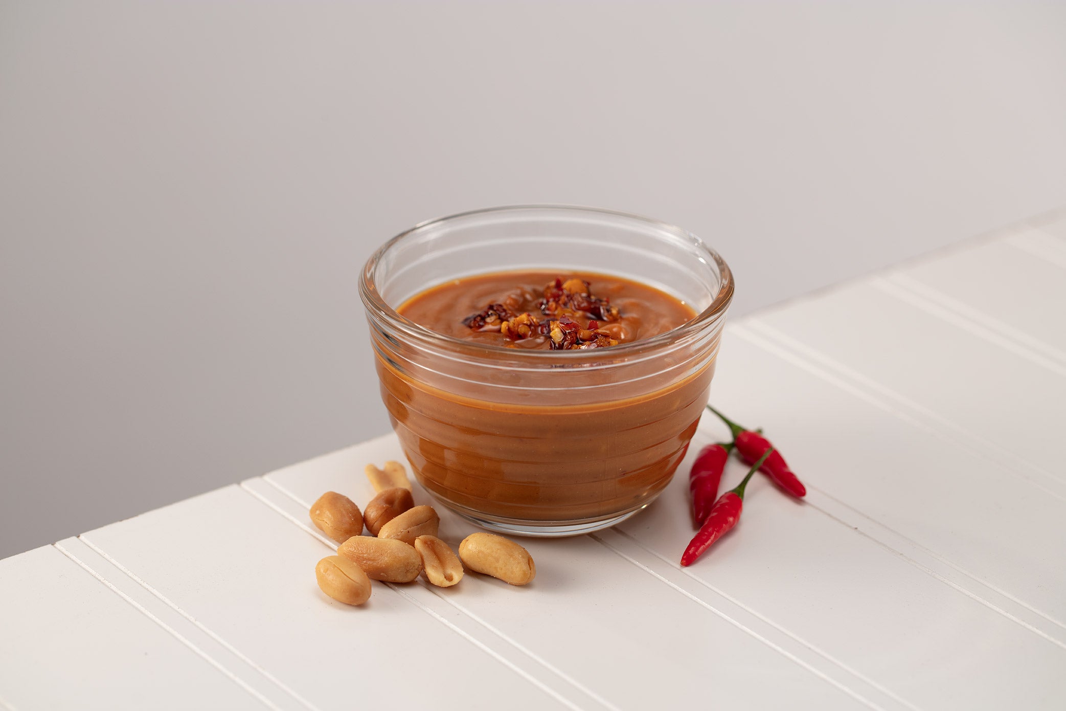 glass bowl with peanut sauce, roasted peanuts and chiles on a white table