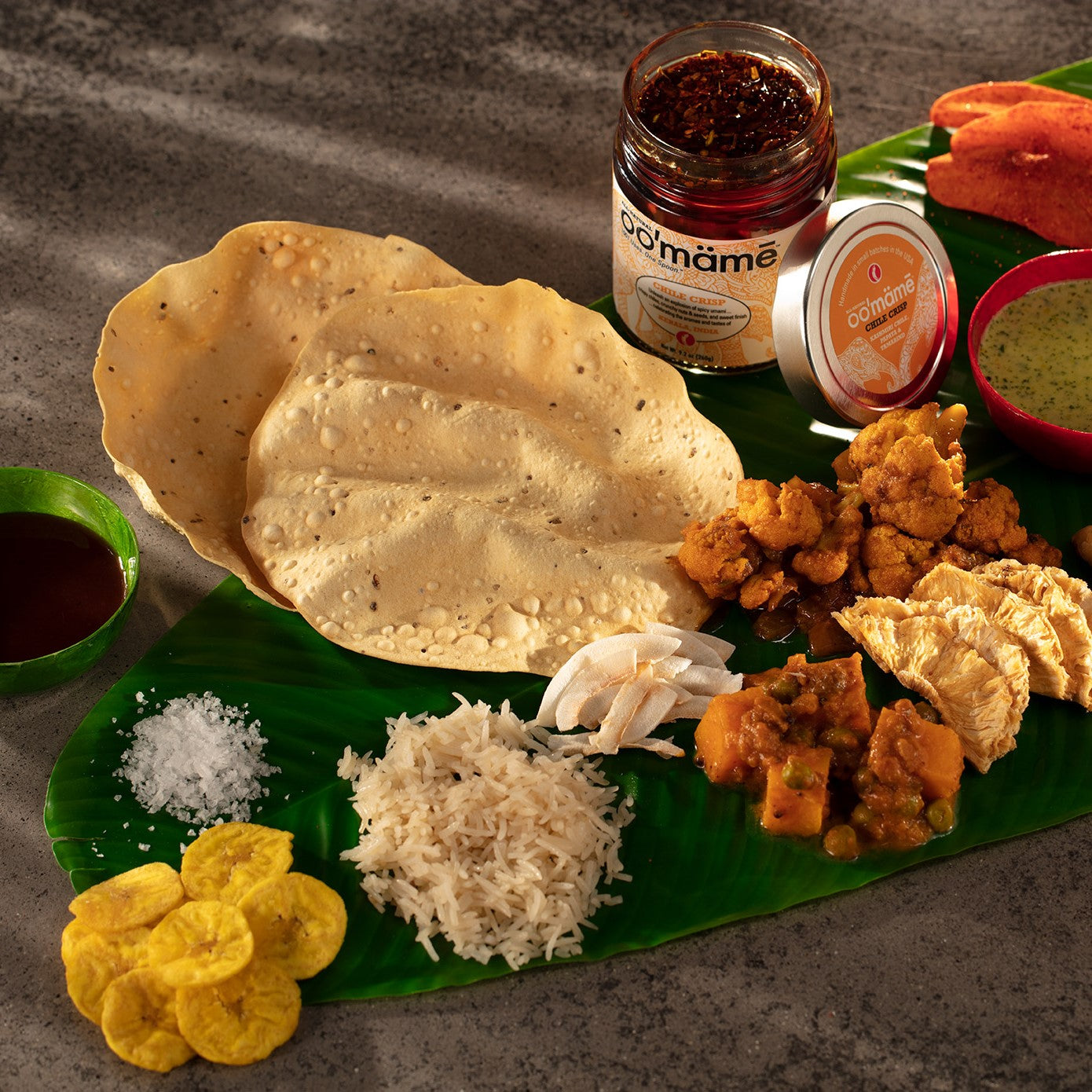 banana leaf covered with Indian vegetarian side dishes, pappadum, chutneys and a jar of oo’mämē Indian Chile Crisp