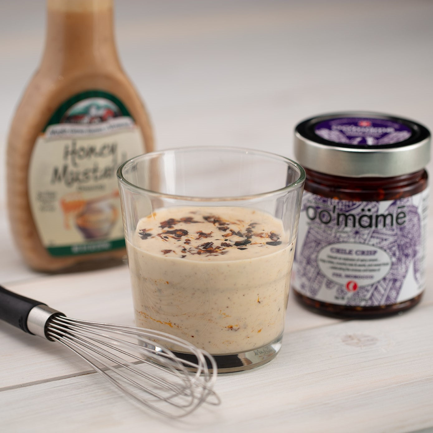 Glass with oo’mämē mixed with bottled salad dressing, jar of Moroccan oo’mämē Chile Crisp, bottle of honey mustard salad dressing and a whisk.