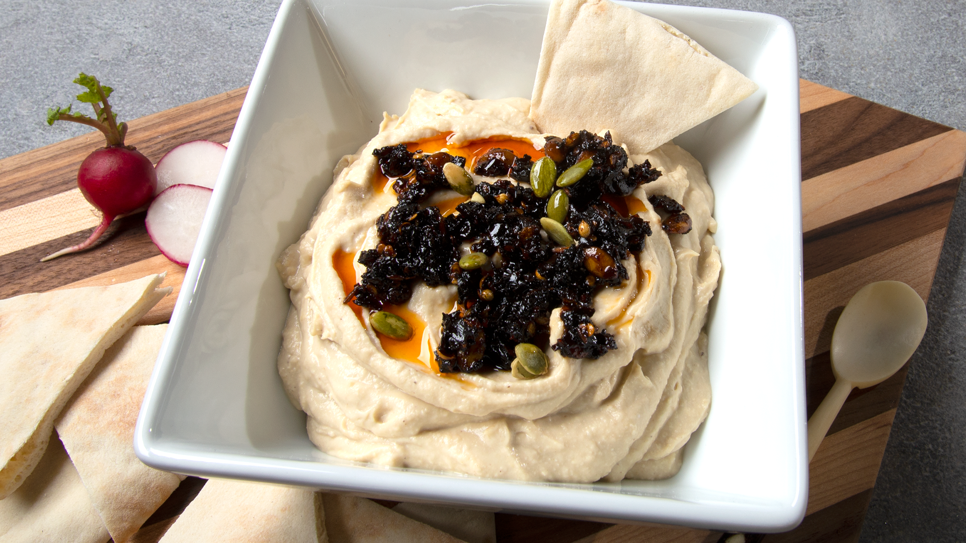 White square bowl with hummus topped with oo’mämē Mexican chili infusion, on a cutting board with pita wedges. #hummus #myoomame