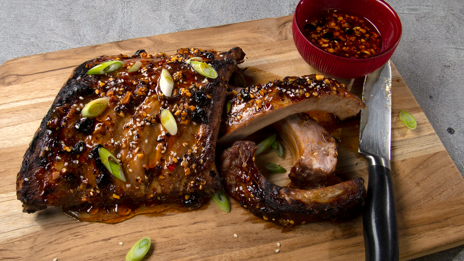 Cutting board with rack of baby back ribs glazed with honey and oo’mämē, a knife and a small red bowl with glaze. #babybackribs #myoomame