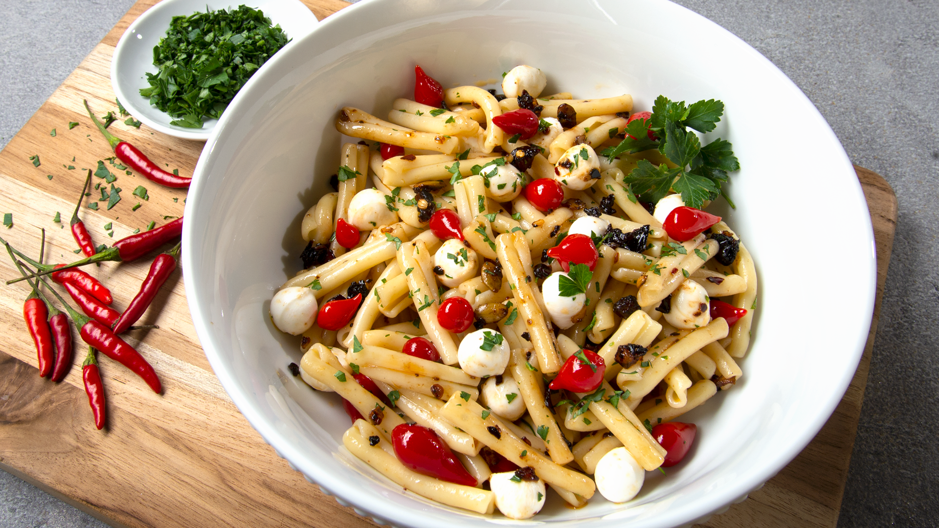 Large white bowl on a cutting board with pasta and mini mozzarella, pickled peppers and oo’mämē. Small saucer of chopped parsley, #spicypasta #myoomame