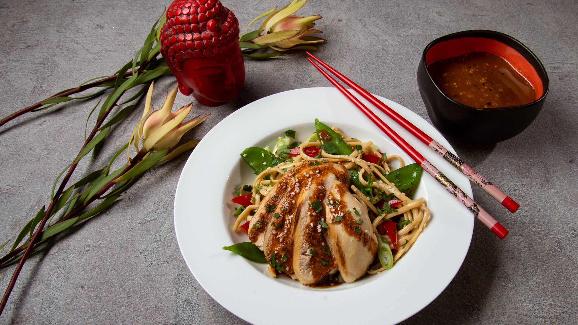 white bowl with noodles, chicken, snow peas, oo’mämē peanut sauce, with chopsticks and flower. Small red Buddha head and black dish with extra oo’mämē peanut sauce 