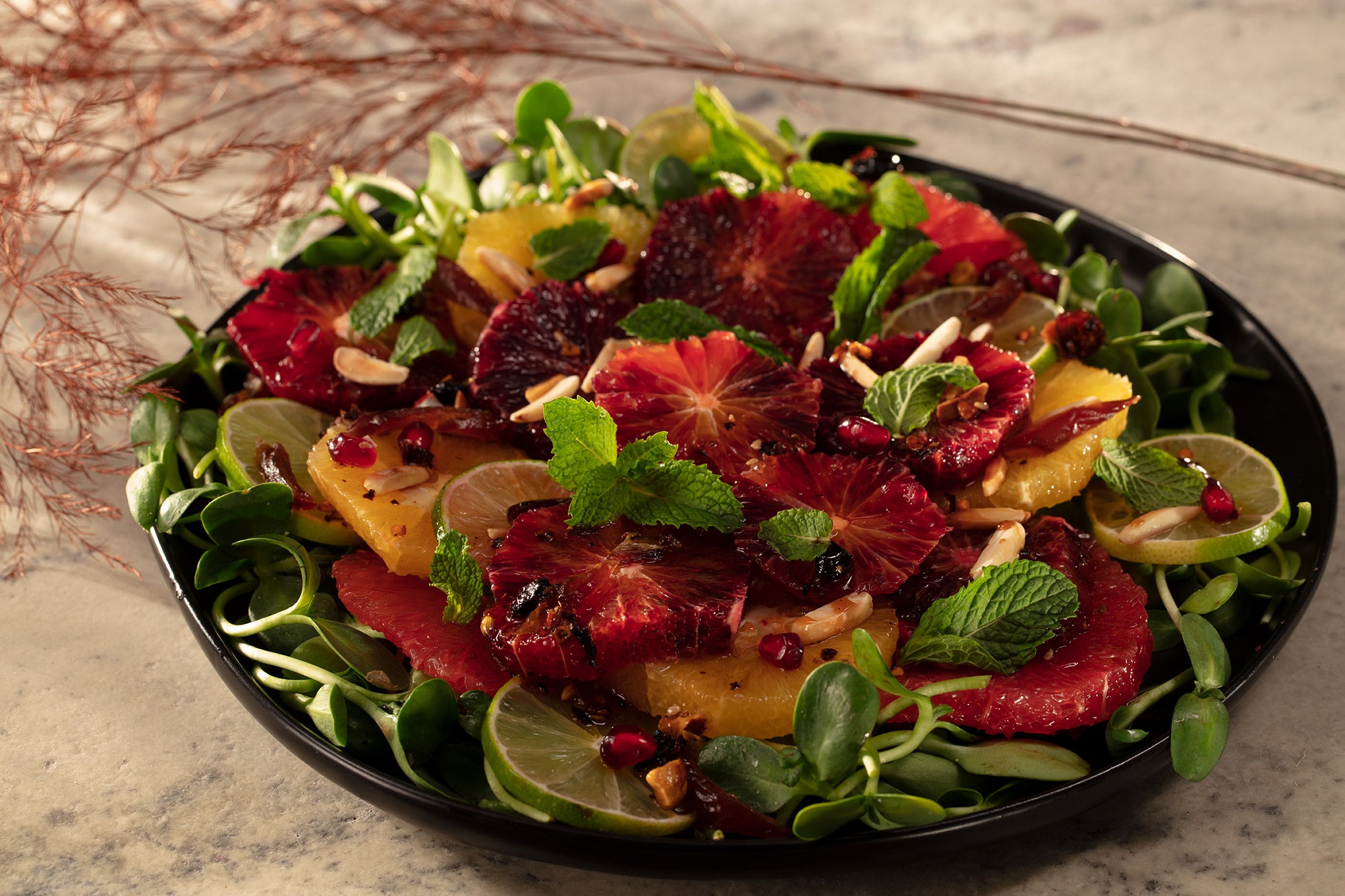 Black plate with bed of sunflower sprouts, topped with sliced navel oranges, blood oranges, red grapefruit and limes. Garnished with almonds, mint leaves, dates and pomegranate arils.