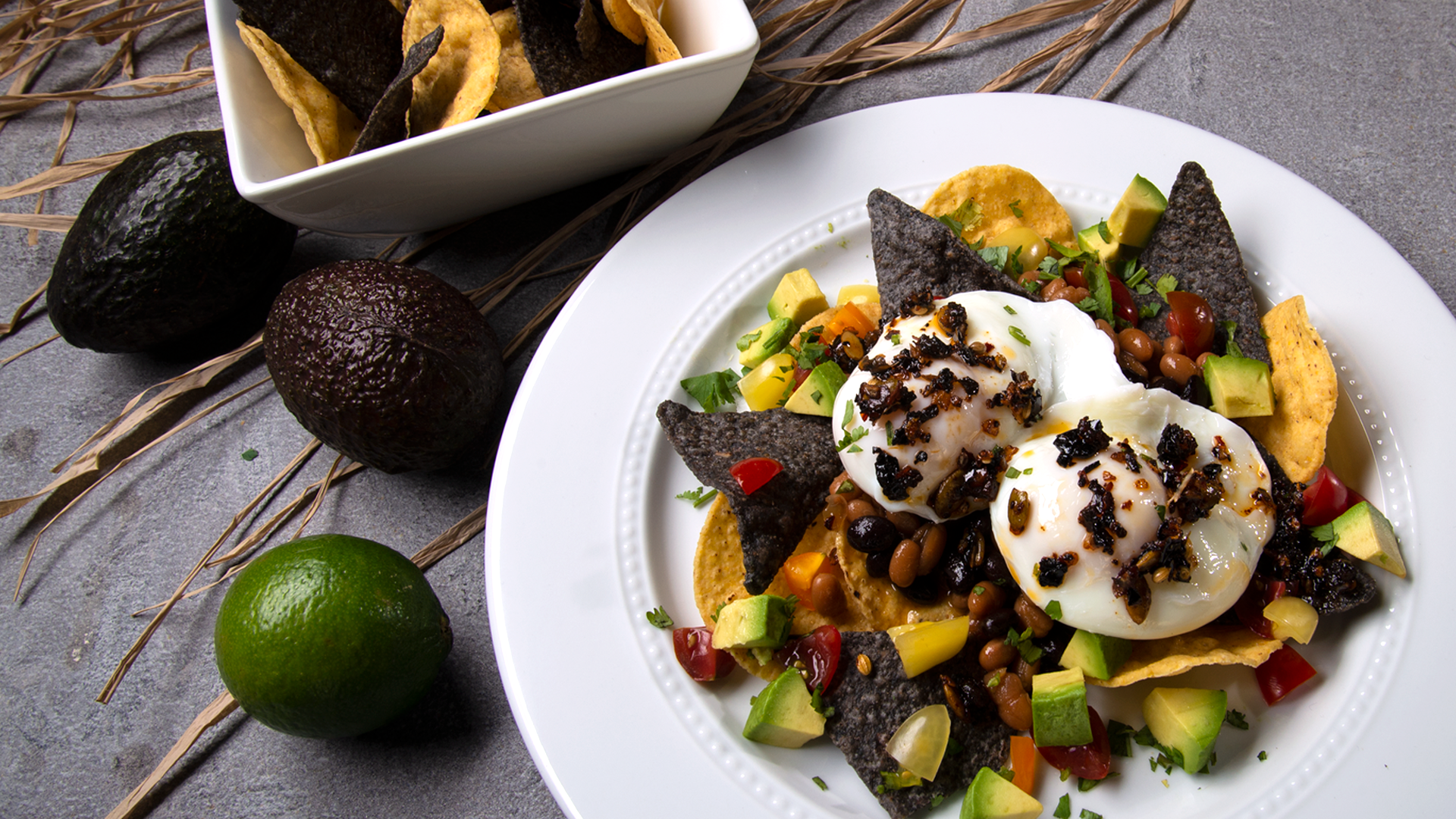 White round plate with chips, poached eggs, topped with Mexican oo’mämē Chile Crisp, avocado and tomatoes. White square bowl with chips and whole avocadoes and lime.