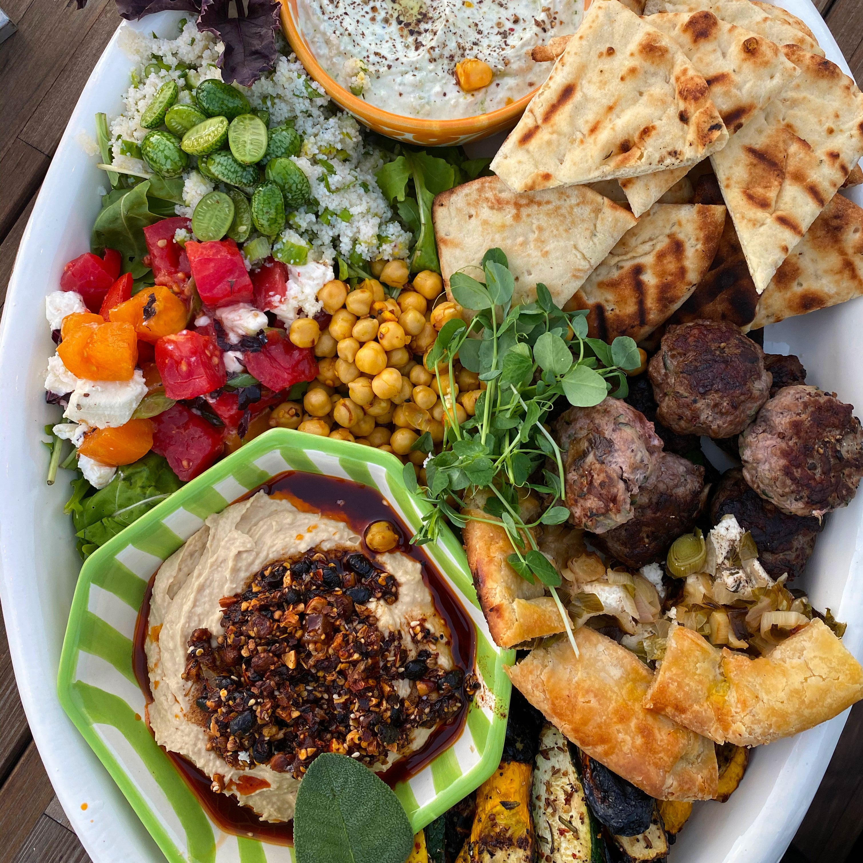 Large white oval platter with oo’mämē lamb sliders, leek galette, grilled squash, hummos topped with oo’mämē, tomato and sheep cheese, fonio with baby cucumbers, garbanzos, naan wedges, yogurt dip with spices and herb garnish.