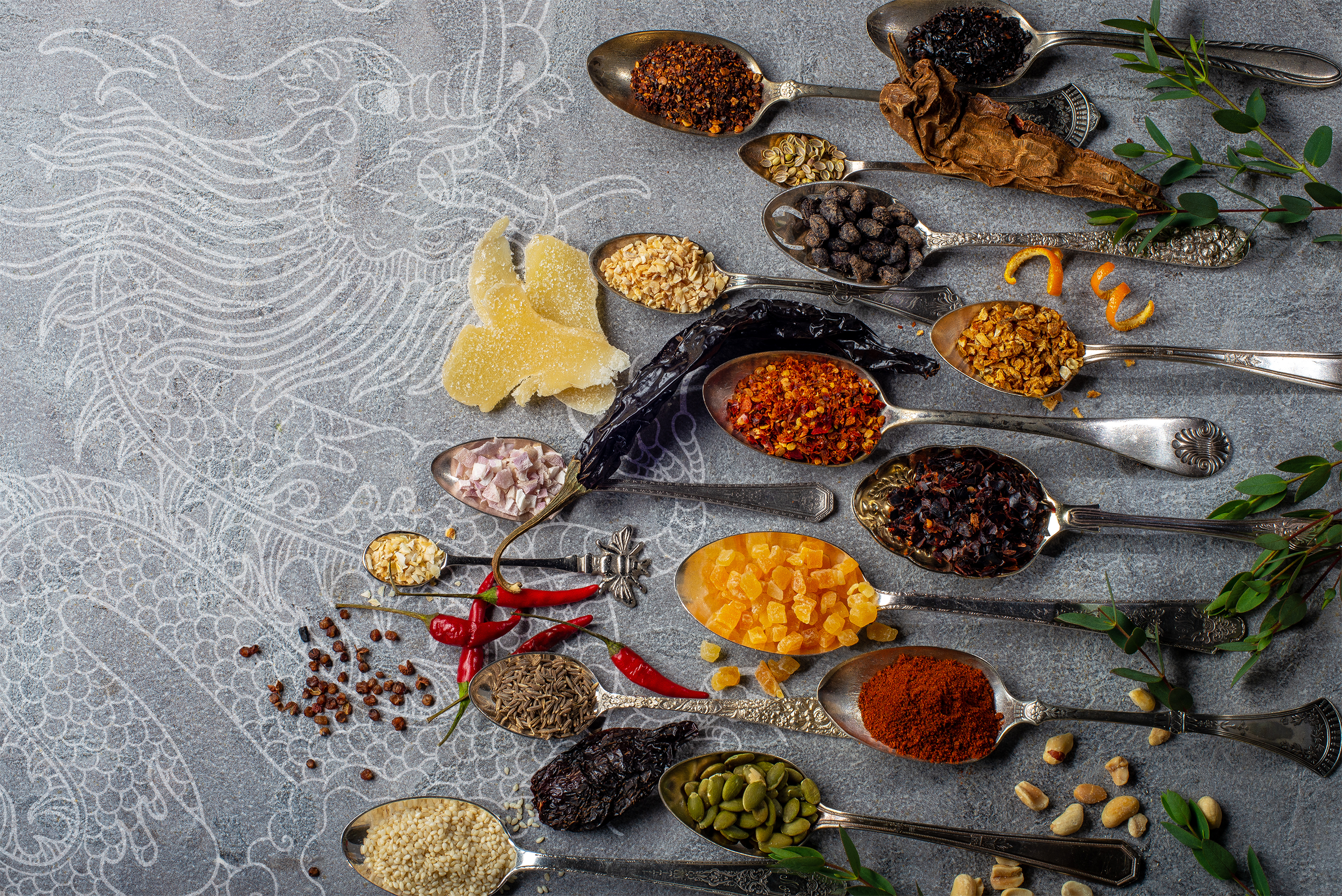 Vintage spoons filled with spices, seeds, chiles and fruits that are ingredients for oo’mämē chile crisp on slate grey background