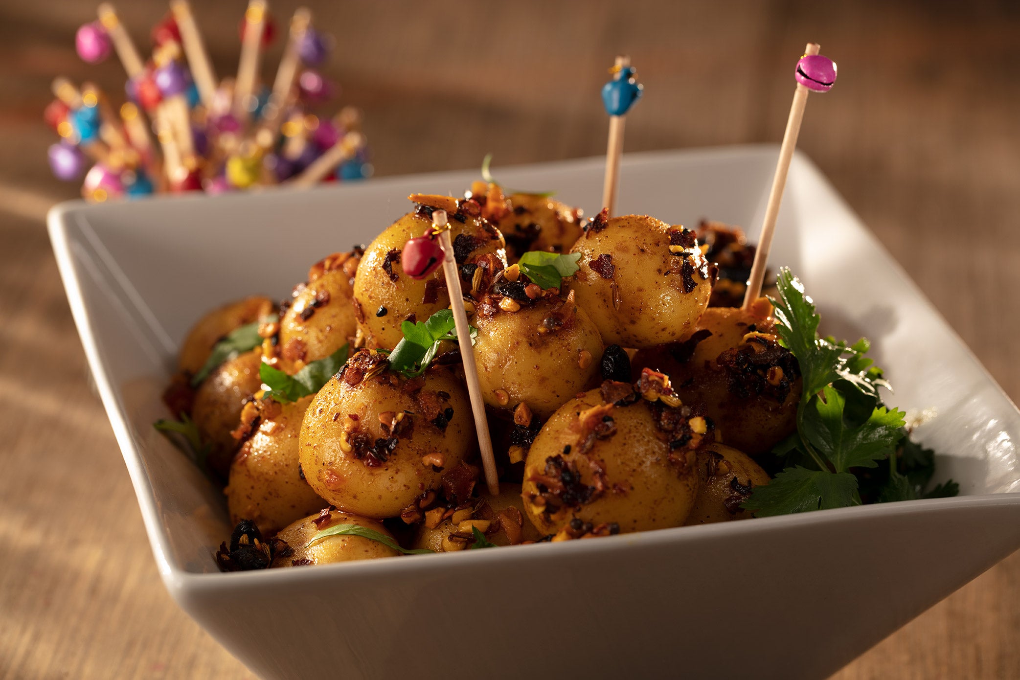Square white bowl with small white potatoes roasted in oo’mämē Indian chile crisp. Container with toothpicks topped with colorful bells.