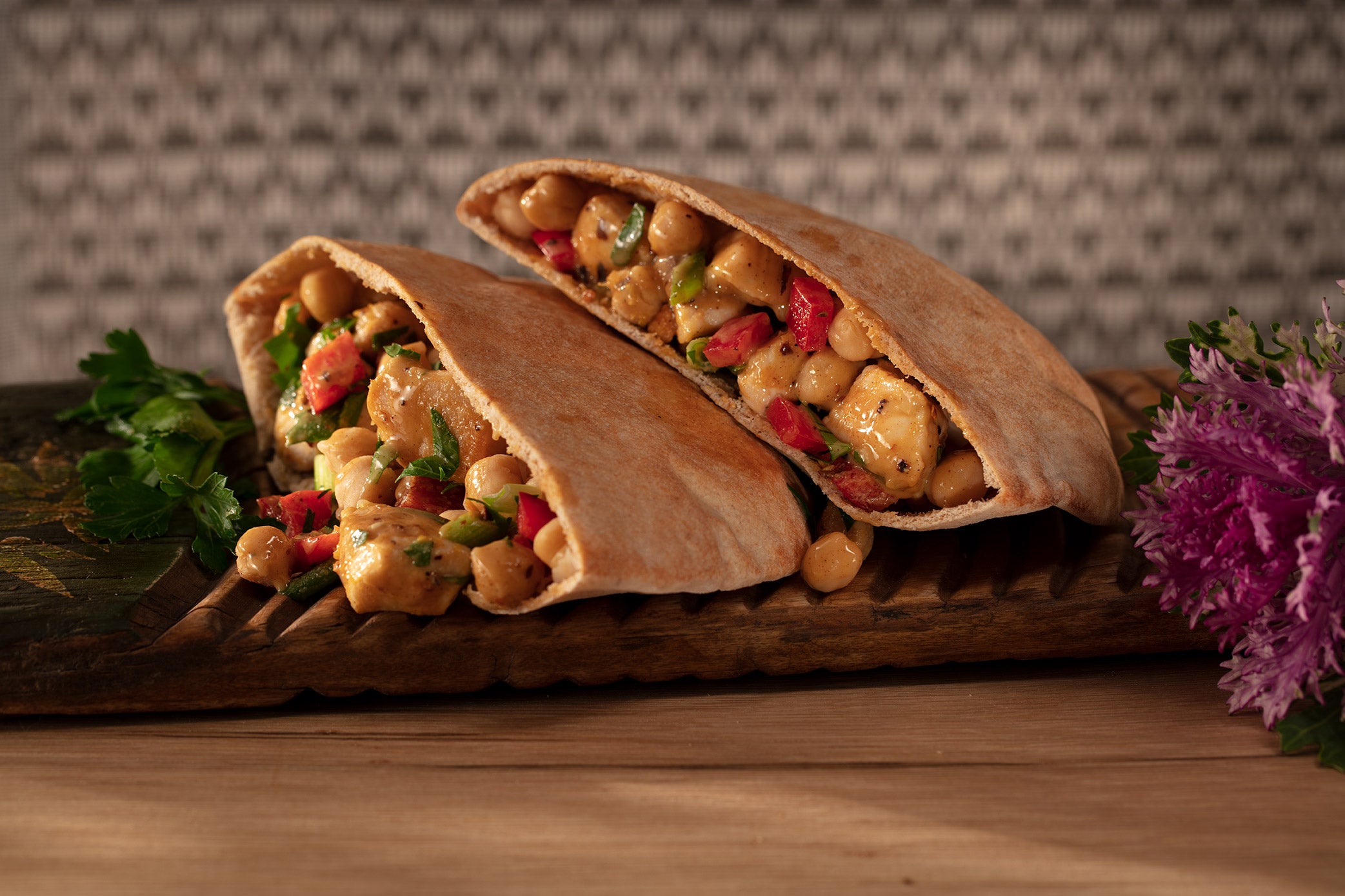 Pita pockets on a cutting board filled with chicken, garbanzo beans, scallions and red peppers with geometric background and flowering kale accent