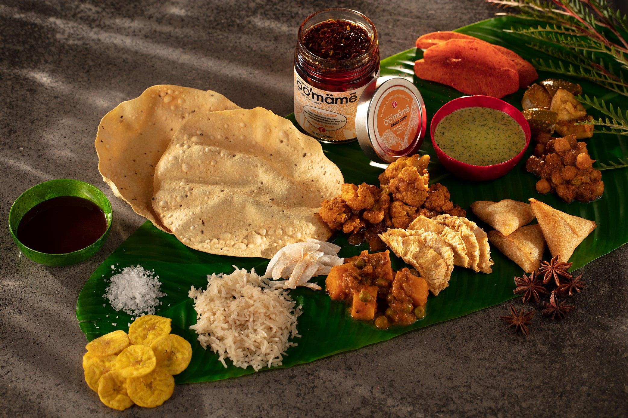 Banana leaf topped with traditional Sadhya components, including pappadum, tamarind and green chutneys in small bowls, jar of oo’mämē Indian chile crisp, banana chips, rice, coconut, pumpkin and peas, curried cauliflower, dried pineapple, samosas, chana, mango achar, chile dusted dried mango