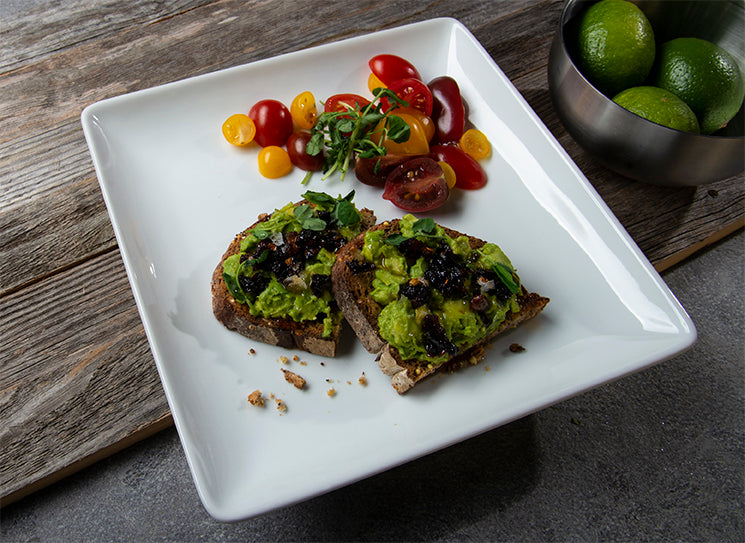 avocado toast with umami oaxacan chili crisp presented on white square plate with tomato garnish and limes in a bowl