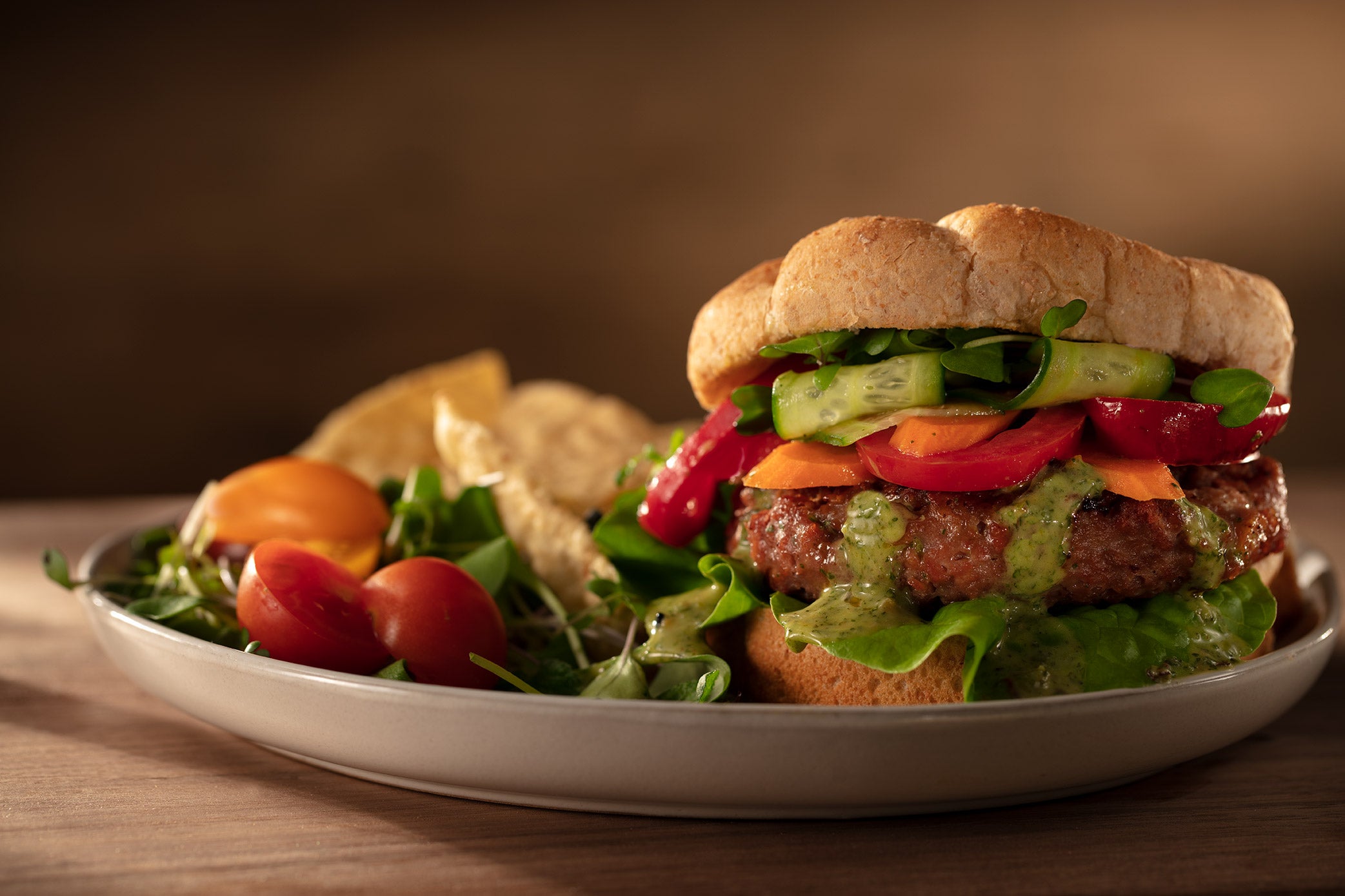 Veggie burger with a bun, green sauce and garnished with lettuce, carrots, tomatoes, cucumbers and sprouts. Garnished with cherry tomatoes and corn chips