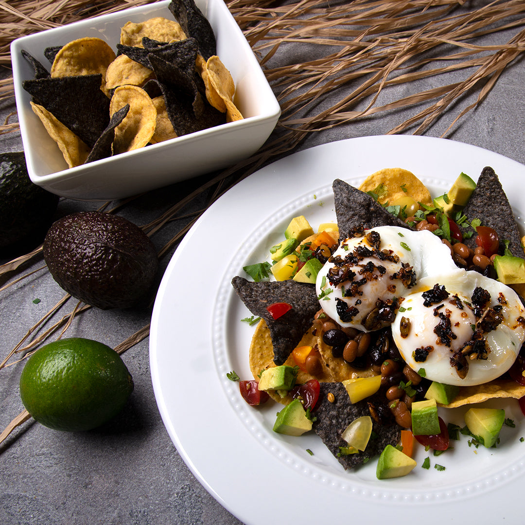 White round plate with chips, poached eggs, topped with Mexican oo’mämē Chile Crisp, avocado and tomatoes #huevos #myoomame  