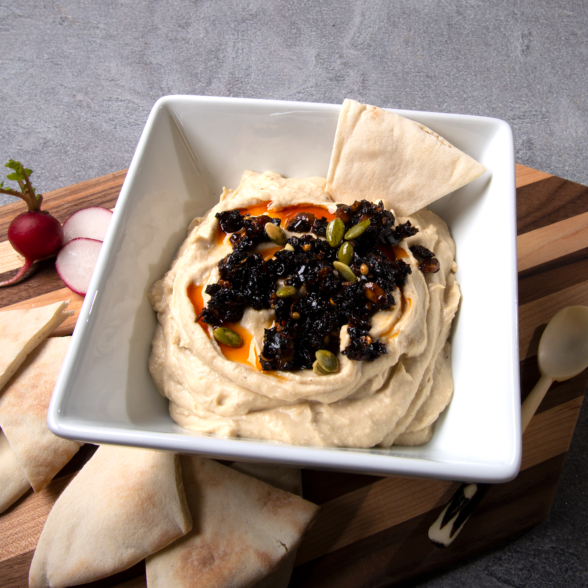 White square bowl with hummus topped with oo’mämē Mexican chile crisp, on a cutting board with pita wedges. #hummus #myoomame
