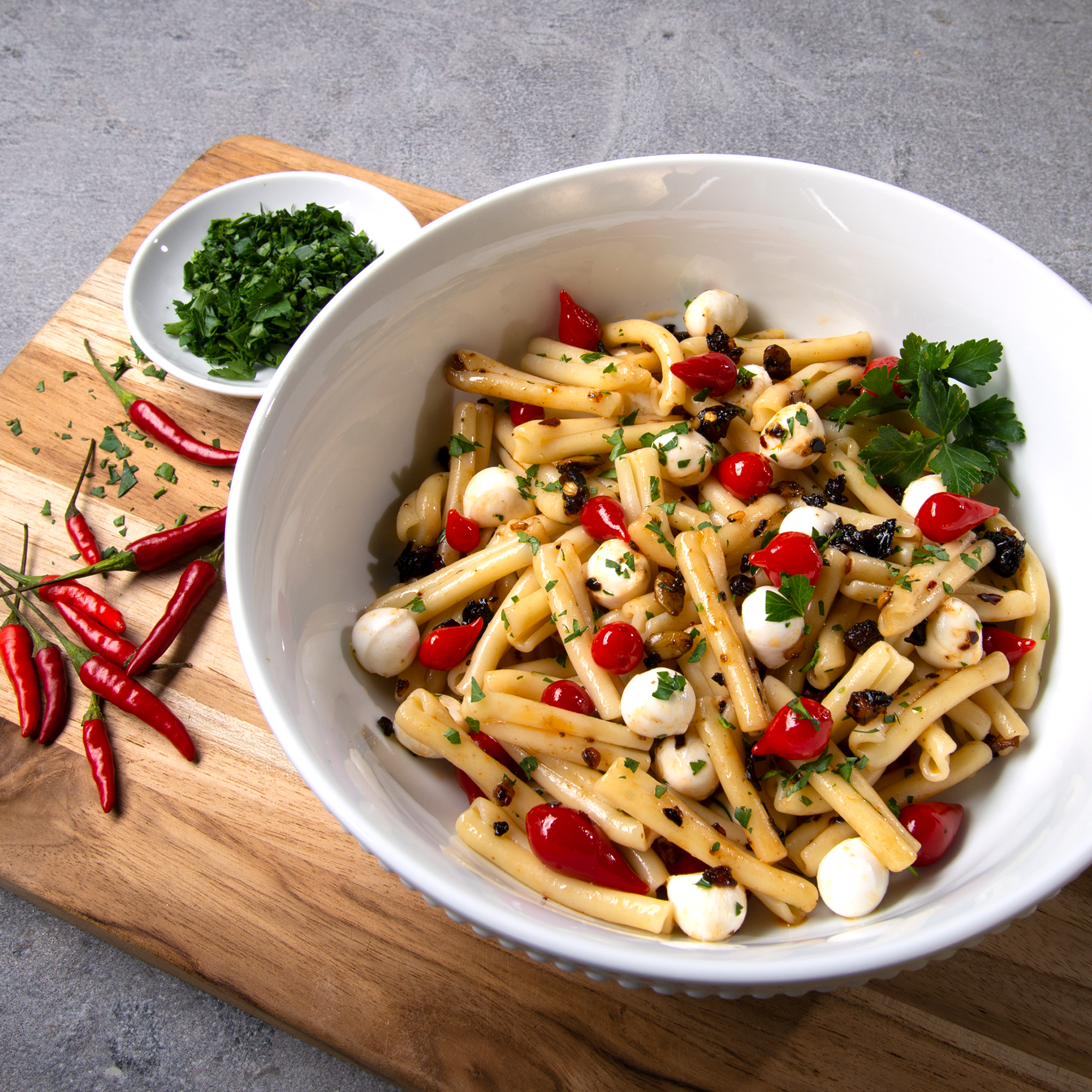 Large white bowl on a cutting board with pasta and mini mozzarella, pickled peppers and oo’mämē. Small saucer of chopped parsley, #spicypasta #myoomame