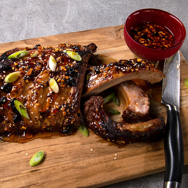 Cutting board with rack of baby back ribs glazed with honey and oo’mämē, a knife and a small red bowl with glaze. #babybackribs #myoomame