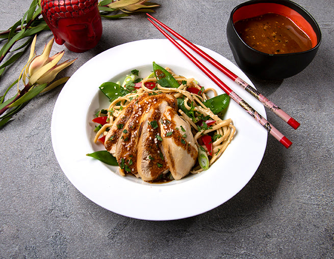 white bowl with noodles, chicken, snow peas, oo’mämē peanut sauce, with chopsticks and flower. Small red Buddha head and black dish with extra oo’mämē peanut sauce 